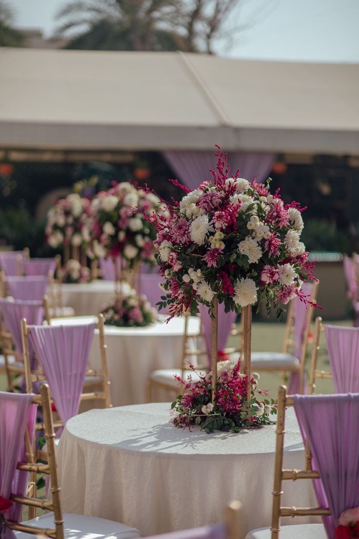 gallery-2 Chic wedding setup with floral centerpiece and lavender chairs outdoors.